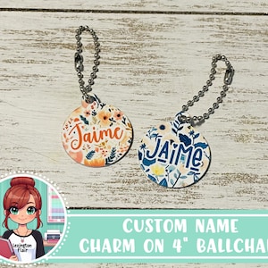 May include: Two round, personalized name charms with floral patterns. The charms are attached to 4 inch silver ball chains. The charms say "Jaime" in a colorful, handwritten font.