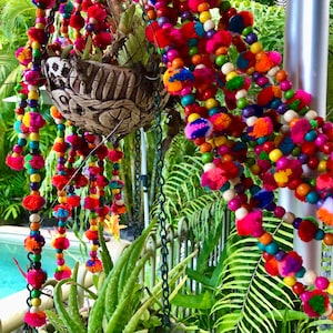 Puede incluir: Una guirnalda colorida de borlas de cuentas con pompones en tonos de rosa, rojo, verde, azul y amarillo. La guirnalda cuelga de una maceta y está rodeada de follaje verde.