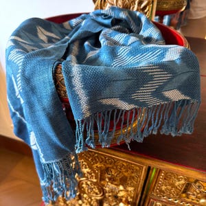 Peut inclure: Écharpe bleue et blanche à motifs avec franges. L'écharpe est drapée sur un bol décoratif rouge et doré. L'écharpe présente un motif tissé avec un mélange de teintes bleu clair et foncé.