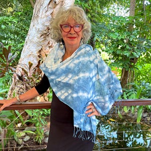May include: A woman wearing a blue and white tie-dye scarf with fringe. She is standing in front of a tree and a pond.