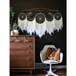 May include: A white dreamcatcher with five hoops and feathers hanging from a wooden branch. The dreamcatcher is hanging on a black wall above a brown dresser and a white chair.