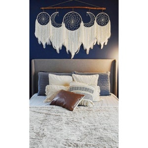 May include: A white and cream colored dreamcatcher with a wooden dowel and fringe hanging above a bed with a white fluffy blanket and pillows. The bed has a gray headboard and a white, cream, and black patterned pillow.