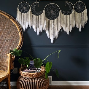 May include: A set of three white and brown dreamcatchers with feathers hanging from a branch, mounted on a dark blue wall. A wicker chair and a wicker side table with a potted plant are in the foreground.