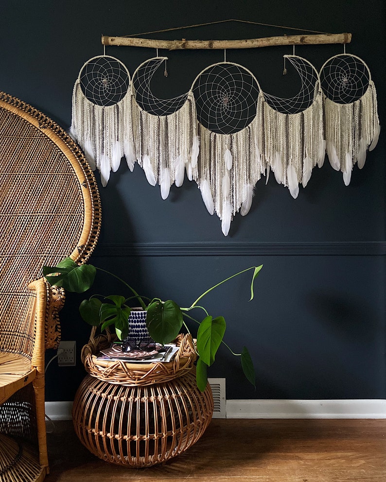 May include: A set of three white and brown dreamcatchers with feathers hanging from a branch, mounted on a dark blue wall. A wicker chair and a wicker side table with a potted plant are in the foreground.