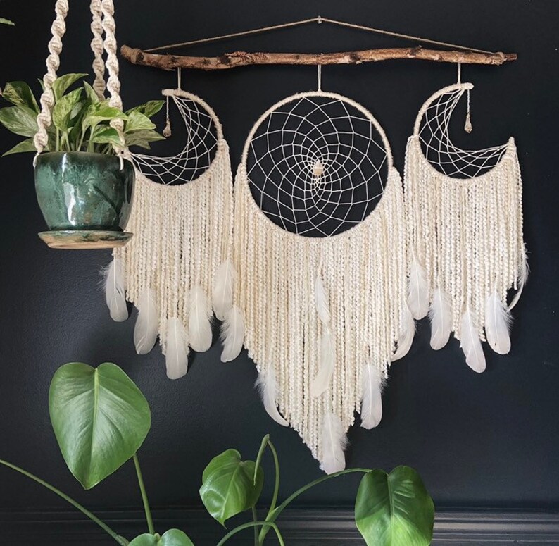 May include: Three white dreamcatchers with feathers hanging from a branch. The dreamcatchers are arranged in a crescent shape. The dreamcatchers are made with white yarn and feathers.