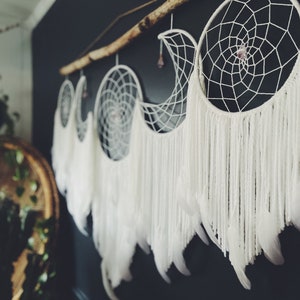 May include: A set of four white dreamcatchers with feathers hanging from a branch. The dreamcatchers are in the shape of a crescent moon and have a small crystal in the center.