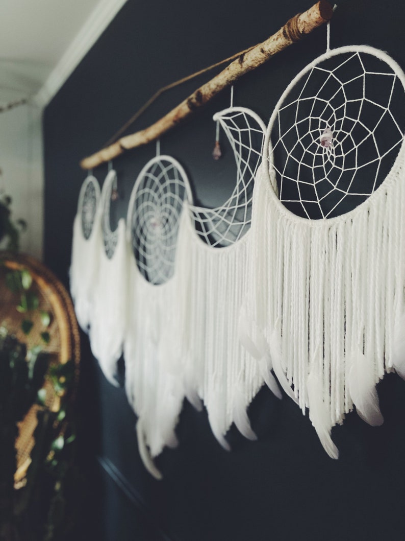 May include: A set of four white dreamcatchers with feathers hanging from a branch. The dreamcatchers are in the shape of a crescent moon and have a small crystal in the center.