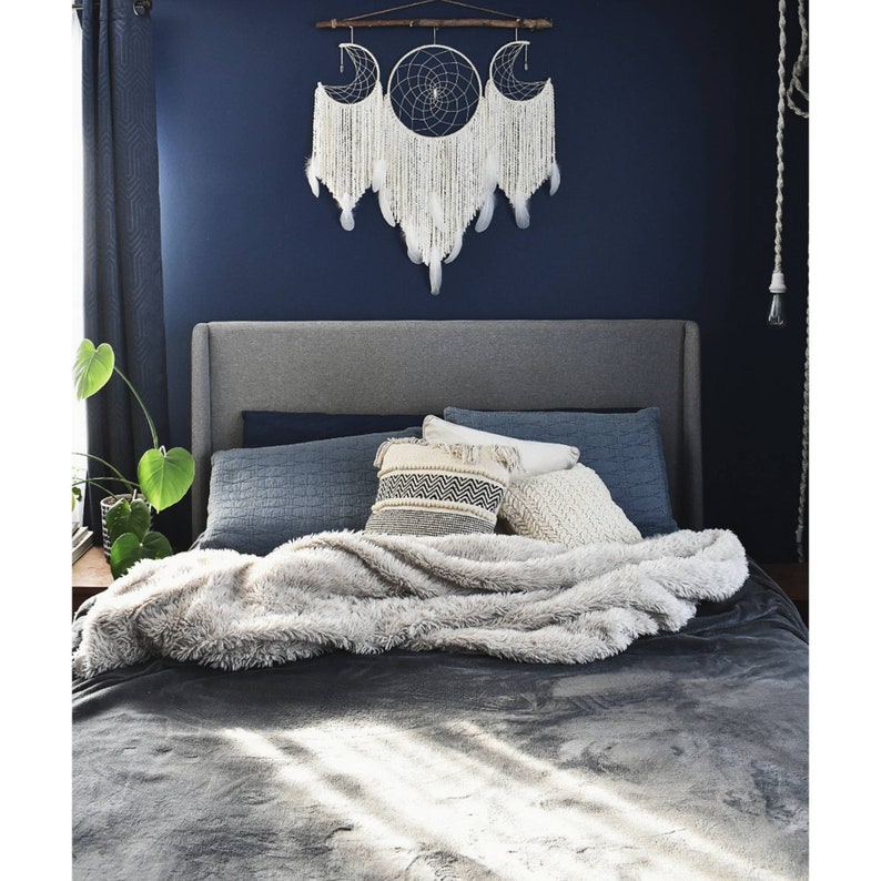 May include: A gray upholstered bed with a white fluffy blanket and a variety of pillows. The headboard is gray and the bed is against a dark blue wall. A white dreamcatcher with feathers hangs above the bed.