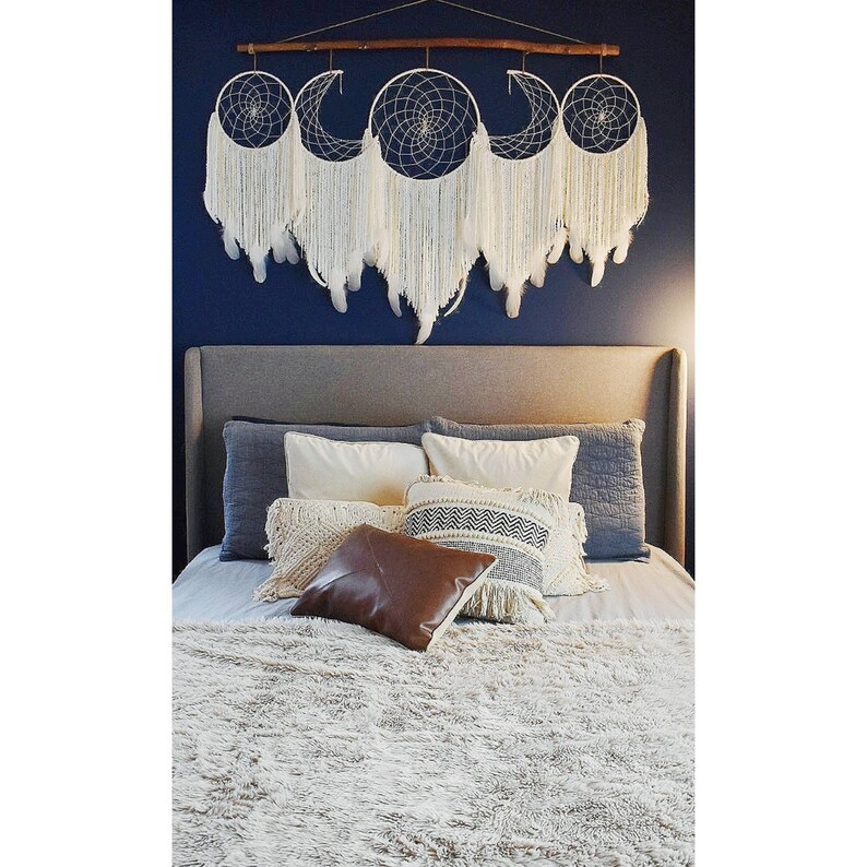 May include: A white and cream colored dreamcatcher with a wooden dowel and fringe hanging above a bed with a white fluffy blanket and pillows. The bed has a gray headboard and a white, cream, and black patterned pillow.