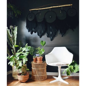 May include: A black and white room with a black feather dreamcatcher hanging on the wall. There are plants in pots on a wicker table and a white chair with a white cushion.