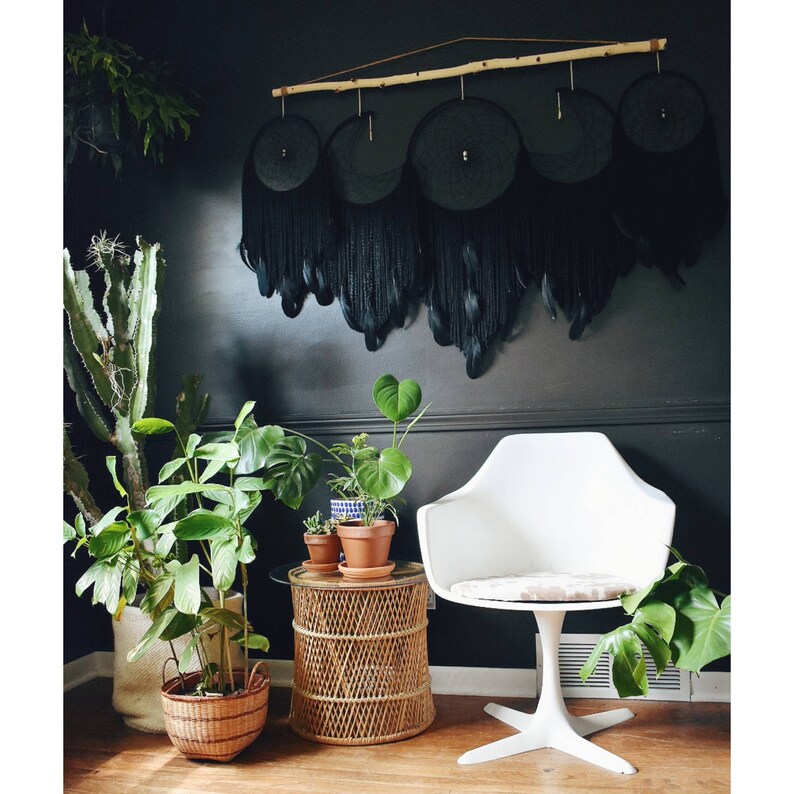 May include: A black and white room with a black feather dreamcatcher hanging on the wall. There are plants in pots on a wicker table and a white chair with a white cushion.