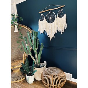 May include: A white macrame dream catcher with three crescent moon shapes hangs on a dark blue wall. The dream catcher is decorated with feathers and beads. A wicker chair and potted plants are in the foreground.