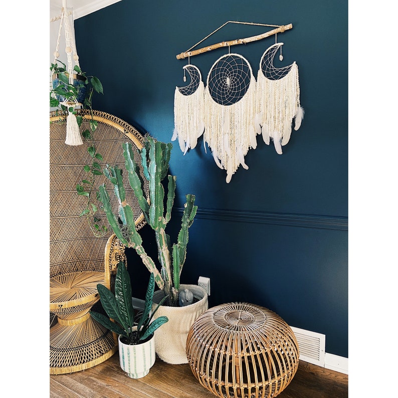 May include: A white macrame dream catcher with three crescent moon shapes hangs on a dark blue wall. The dream catcher is decorated with feathers and beads. A wicker chair and potted plants are in the foreground.