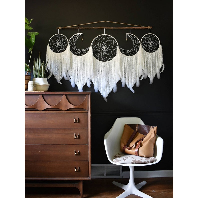 May include: A white dreamcatcher with five hoops and feathers hanging from a wooden branch. The dreamcatcher is hanging on a black wall above a brown dresser and a white chair.
