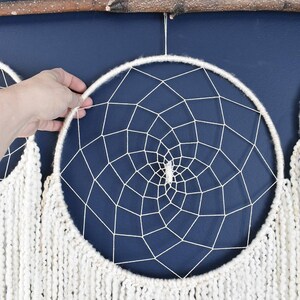 May include: Two white dreamcatchers with white yarn fringe hanging from a branch against a dark blue wall. The dreamcatchers have a white web design and a white crystal in the center.