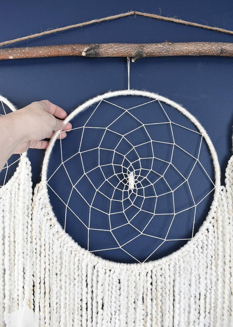 May include: Two white dreamcatchers with white yarn fringe hanging from a branch against a dark blue wall. The dreamcatchers have a white web design and a white crystal in the center.