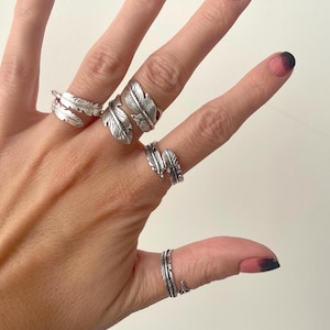 May include: A hand shows off several silver feather rings. The rings have different designs, some wrapping around the finger and others with a single feather. The rings are worn on various fingers, highlighting their different styles.