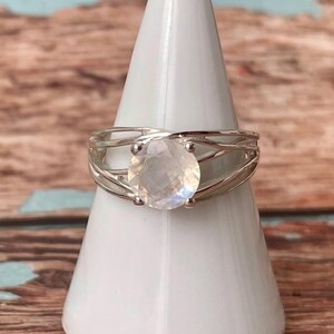 Op de afbeelding: Een zilveren ring met een grote, ronde, iriserende maansteen. De band van de ring heeft een geweven ontwerp. De ring wordt getoond op een witte, kegelvormige standaard tegen een vage houten achtergrond.