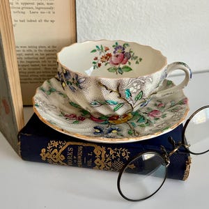 May include: A vintage teacup and saucer set with floral designs in pink, blue, and yellow. The teacup and saucer are placed on a dark blue book, with a pair of round eyeglasses resting nearby. A book is in the background.
