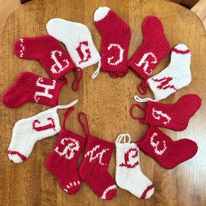 Peut inclure: Une collection de petites chaussettes de Noël tricotées en rouge et blanc. Chaque chaussette présente une lettre différente en blanc, avec une boucle pour suspendre. Les chaussettes sont disposées en cercle sur une surface en bois.