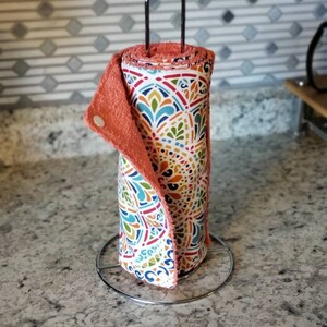 以下が含まれることがあります： 金属製のスタンドに置かれた、カラフルな模様の布製のキッチンタオルロール。タオルは折り畳まれており、コーラル色の裏地が付いています。