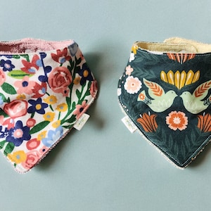 May include: Two baby bibs with different floral patterns. One bib has a pink and white floral pattern with green leaves. The other bib has a dark green background with a floral pattern and two green birds.