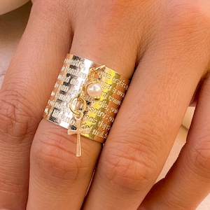 Può includere: Un anello dorato con una fascia larga e una superficie strutturata. L'anello presenta un piccolo ciondolo a forma di croce e una perla. La fascia è incisa con testo. Un gioiello delicato ed elegante.