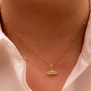 May include: A delicate gold necklace with a small pendant. The necklace features a thin chain with tiny gold beads and a pendant with an intricate design. The necklace is worn over a white collared shirt.