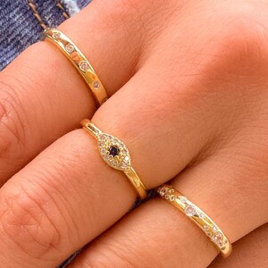May include: Three gold rings with small, clear gemstones. One ring features an oval design with a dark blue center stone. The rings are displayed on a hand with pink nail polish, set against a denim background.
