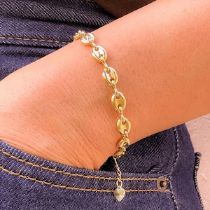 Puede incluir: Una pulsera dorada con un diseño de eslabones de cadena se lleva en la muñeca. La pulsera tiene un pequeño dije de corazón colgando. La persona lleva vaqueros azules y una camiseta marrón.