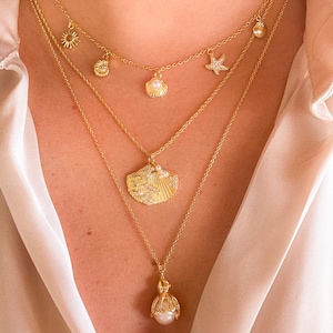 May include: A collection of gold necklaces with ocean-themed pendants. The necklaces feature charms like a sun, starfish, and seashells, and a large shell pendant. The necklaces are layered and worn over a light pink satin shirt.
