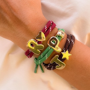 Pode incluir: Close-up de quatro pulseiras com pingentes dourados. As pulseiras são feitas de corda vermelha, verde e marrom. Os pingentes têm a forma das letras R, P e T, e de uma estrela. As pulseiras são usadas no pulso.