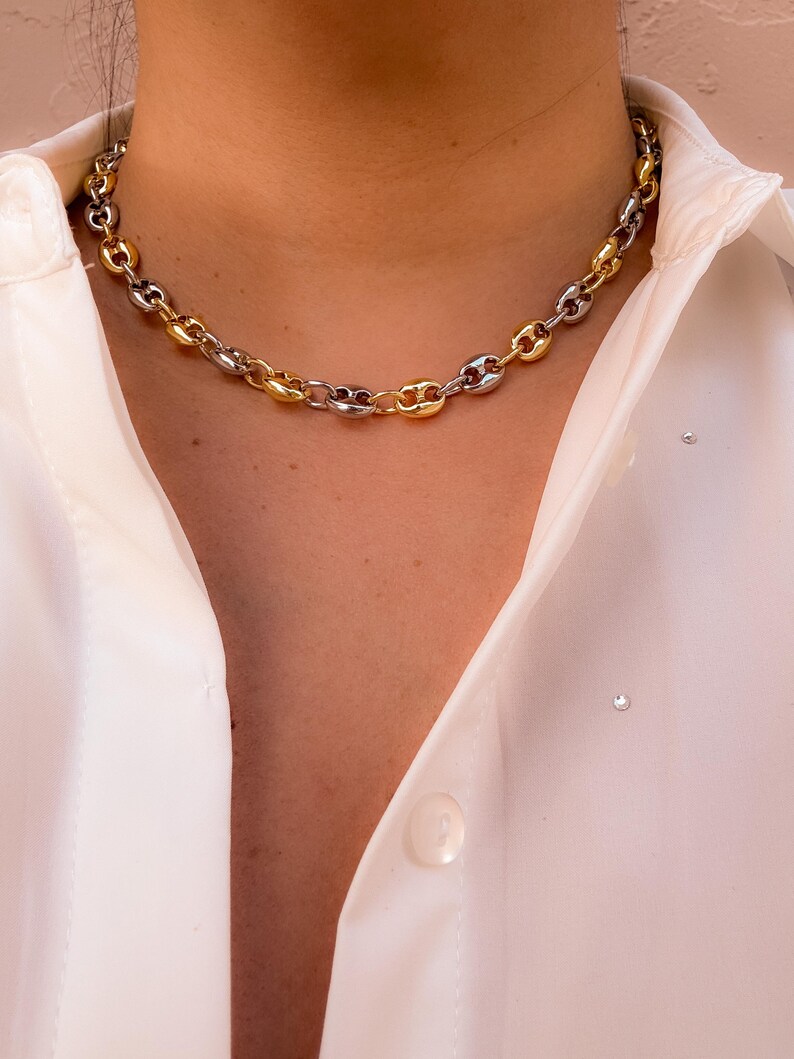 May include: A two-tone chain necklace with alternating gold and silver links. The necklace is a chunky style, with each link resembling a puffed oval. The necklace is worn against a white collared shirt.