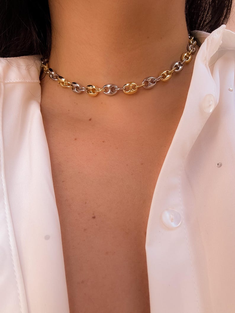 May include: A gold and silver-toned chain necklace with a coffee bean link design. The necklace is worn against a white button-down shirt. The chain alternates between gold and silver links, creating a two-tone effect.