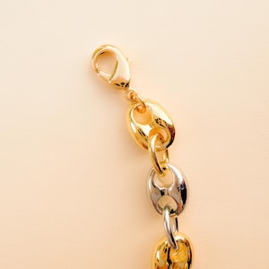 May include: Close-up of a gold and silver chain necklace with a lobster clasp. The chain features a mix of gold and silver links in a puffed, oval shape. The background is a solid, light beige color.