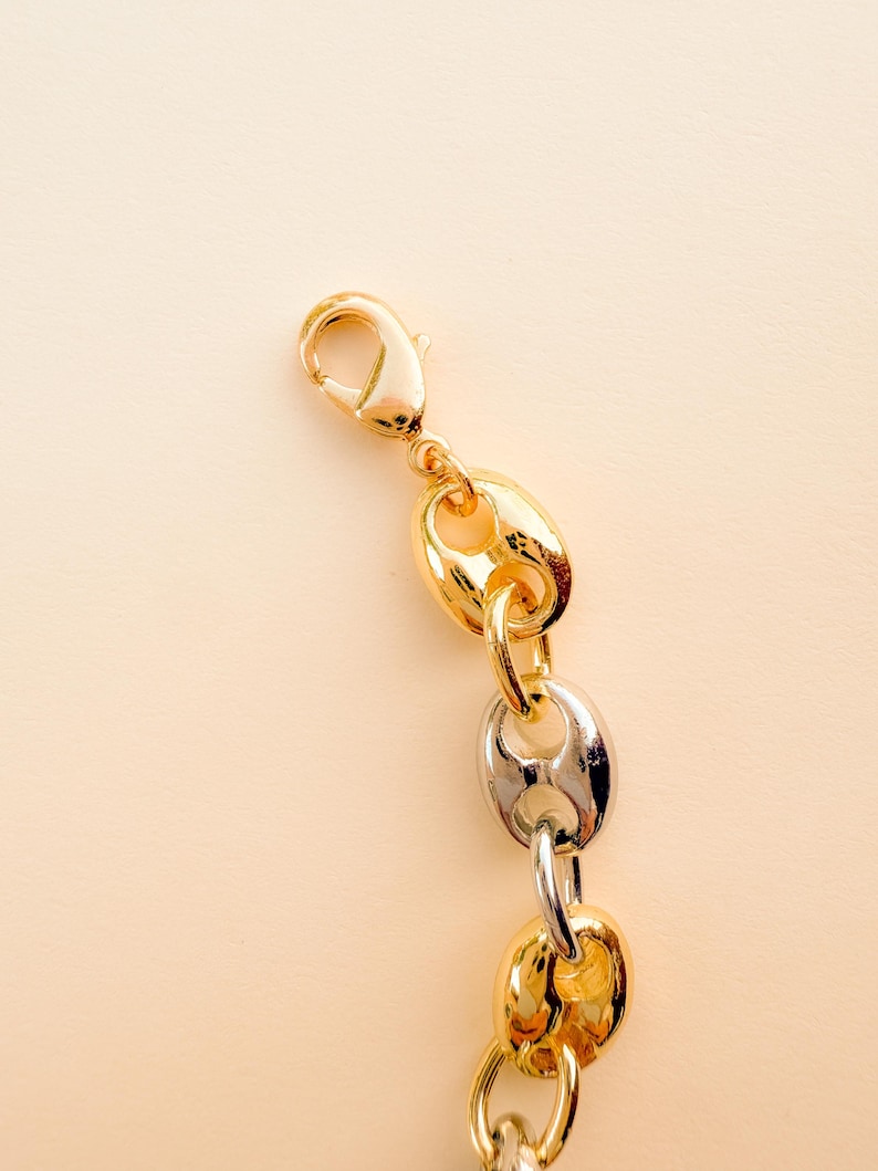 May include: Close-up of a gold and silver chain necklace with a lobster clasp. The chain features a mix of gold and silver links in a puffed, oval shape. The background is a solid, light beige color.