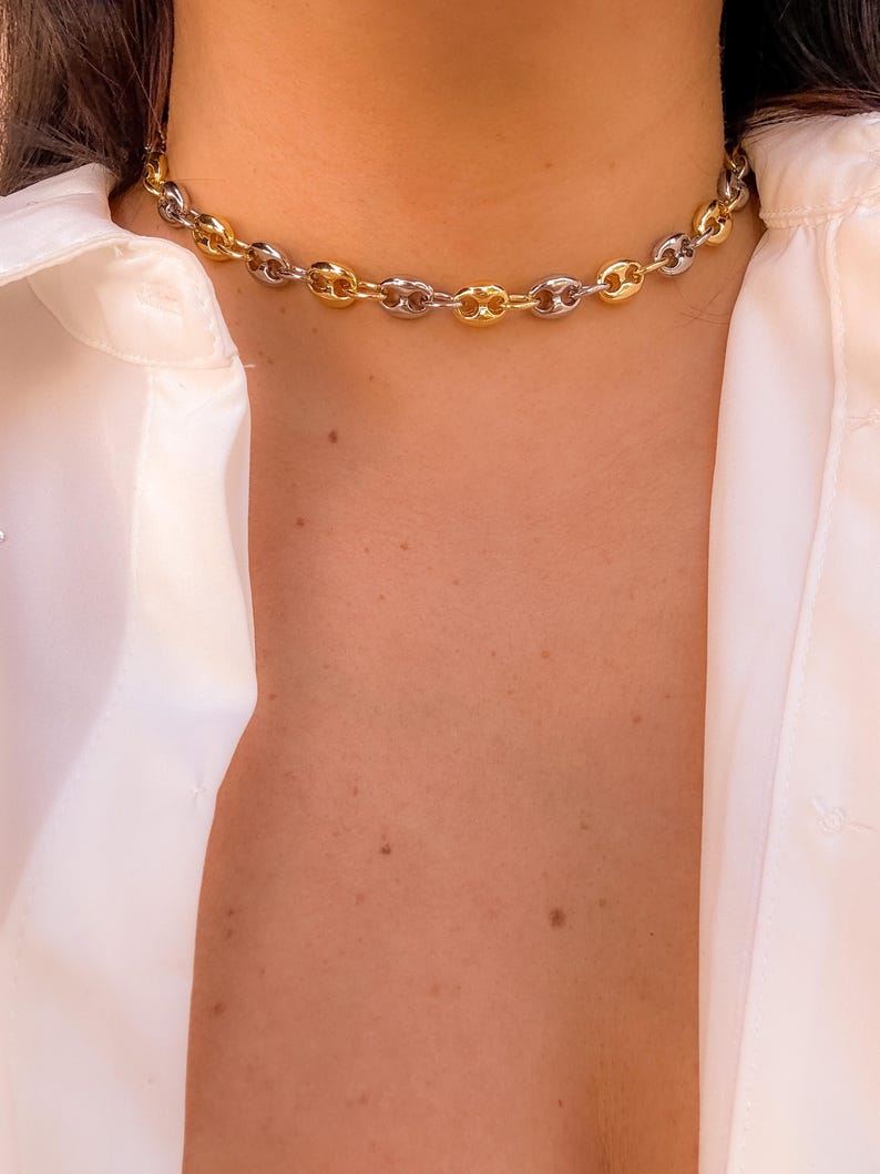 May include: A gold and silver-toned chain necklace with a linked design. The necklace is worn against a person's skin and a white collared shirt.