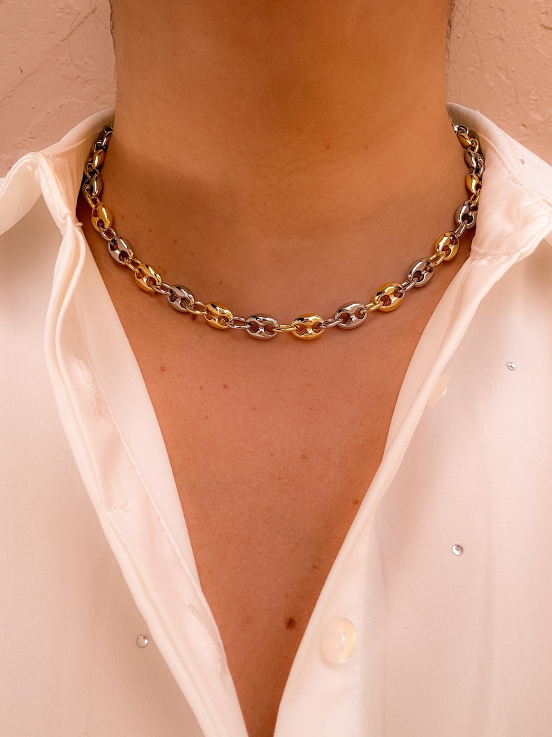 May include: A two-tone chain necklace with gold and silver links. The necklace is made of a series of oval-shaped links, creating a bold and stylish accessory. The necklace is worn over a white collared shirt.