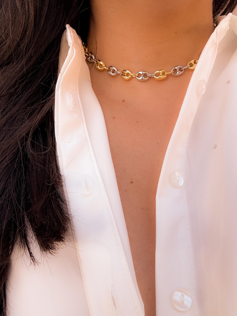 May include: A gold and silver-toned chain necklace with a chunky link design. The necklace is worn against a white button-down shirt. The necklace is a choker style and is a mix of gold and silver links.