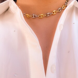 May include: A gold and silver-toned chain necklace with a bold, linked design. The necklace is worn against a white button-down shirt. The chain links alternate between gold and silver, creating a striking contrast. The shirt has small, sparkling embellishments.