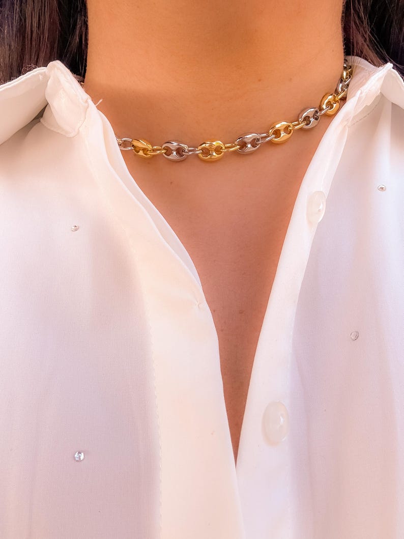May include: A gold and silver-toned chain necklace with a bold, linked design. The necklace is worn against a white button-down shirt. The chain links alternate between gold and silver, creating a striking contrast. The shirt has small, sparkling embellishments.