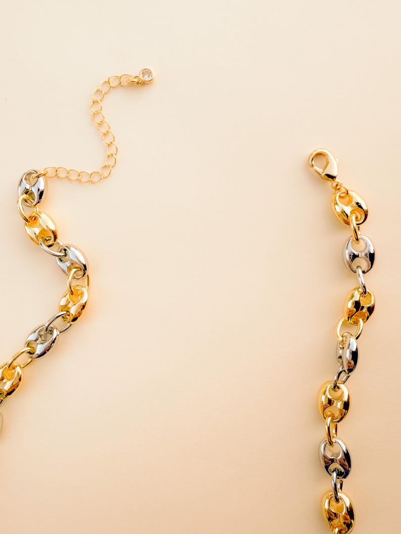 May include: A gold and silver-toned chain necklace with a lobster clasp and an adjustable chain. The necklace features a series of oval-shaped links in alternating gold and silver colors. A small, clear gem is set at the end of the adjustable chain.