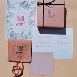 May include: A flat lay of jewelry packaging and accessories. The items include a pink box with a black ribbon, a pink pouch, a cleaning cloth, and a card with the text "RARE pearl jewels". The background is a light peach color.