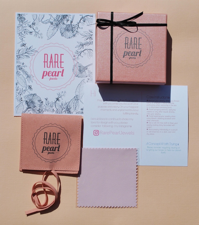 May include: A flat lay of jewelry packaging and accessories. The items include a pink box with a black ribbon, a pink pouch, a cleaning cloth, and a card with the text "RARE pearl jewels". The background is a light peach color.