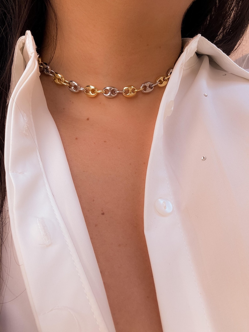 May include: A two-tone chain necklace with alternating gold and silver links. The necklace is a choker style, resting close to the neck. The links are a rounded, puffed design. The necklace is worn over a white button-down shirt.