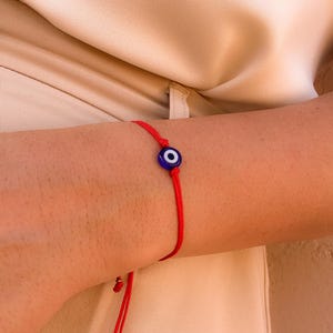May include: A red string bracelet with a blue and white evil eye charm. The bracelet is tied with a sliding knot for adjustable sizing. The charm is a dark blue circle with a white and black center.