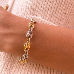 May include: A mixed-metal bracelet featuring a chain link design with alternating gold and silver-toned links. The bracelet is worn on a wrist, with a cream-colored sweater in the background.