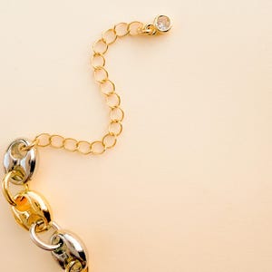 May include: A gold and silver chain necklace with a clear crystal charm. The chain features a mix of gold and silver links, with a delicate gold chain extending to a crystal charm. The background is a solid, light beige color.
