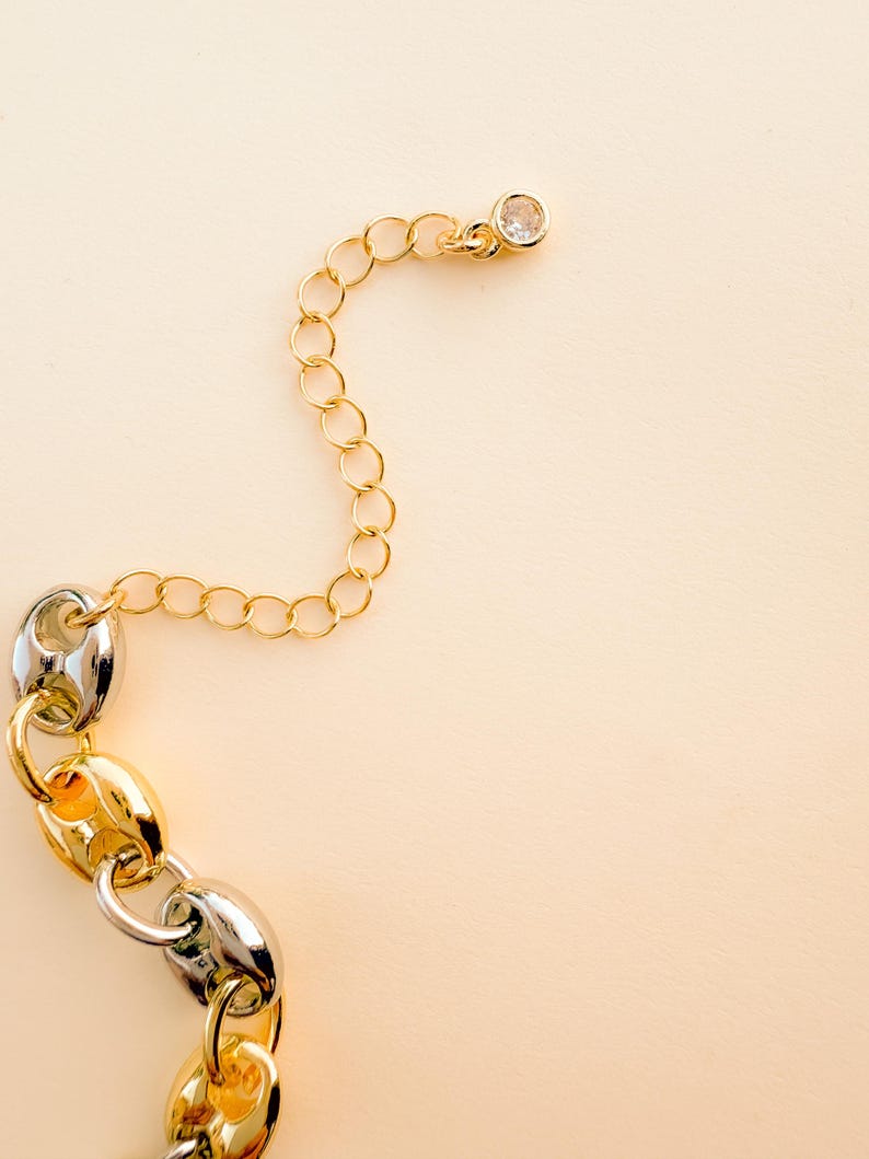 May include: A gold and silver chain necklace with a clear crystal charm. The chain features a mix of gold and silver links, with a delicate gold chain extending to a crystal charm. The background is a solid, light beige color.
