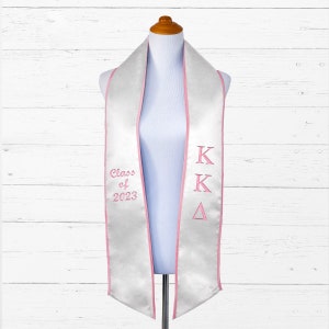 May include: White graduation stole with pink trim and the letters "KK".  The stole also has the text "Class of 2023" embroidered on it.